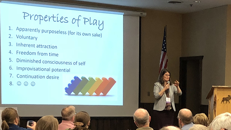 A woman presenting at a conference with a PowerPoint slide titled Properties of Play.