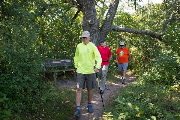 Three people walking on a narrow dirt trail toward the camera during summer.