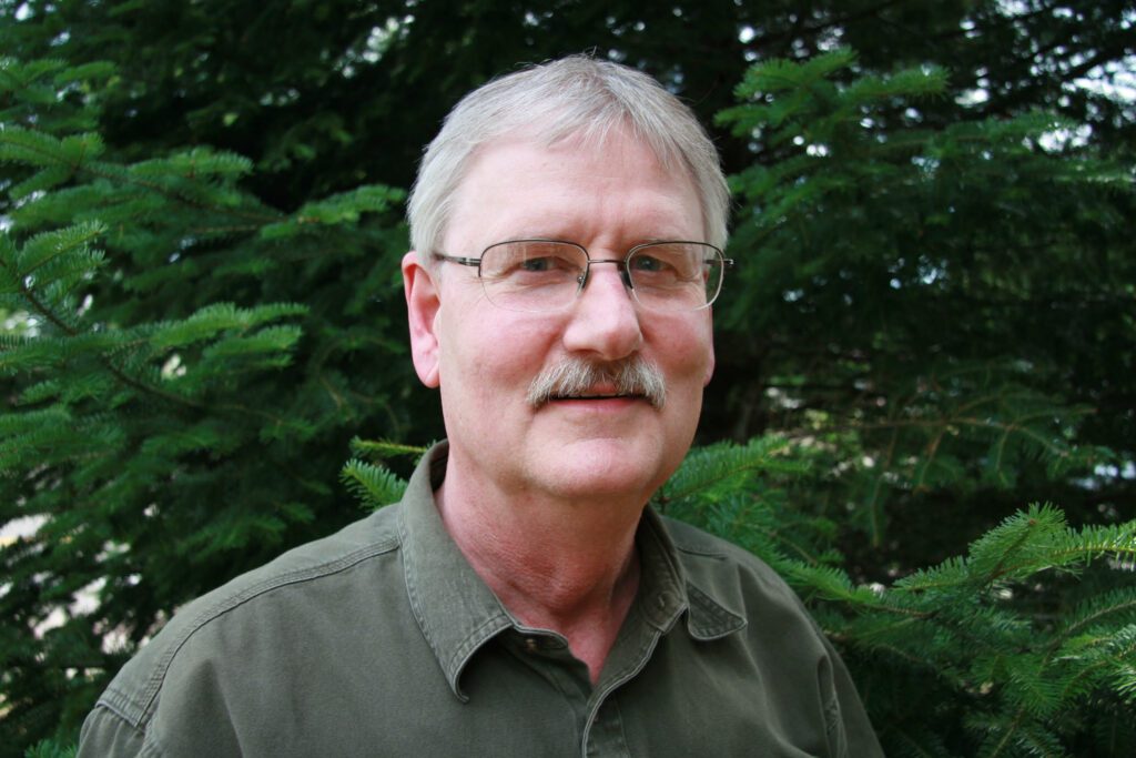 An older white man with glasses and a mustache standing outside in front of trees.