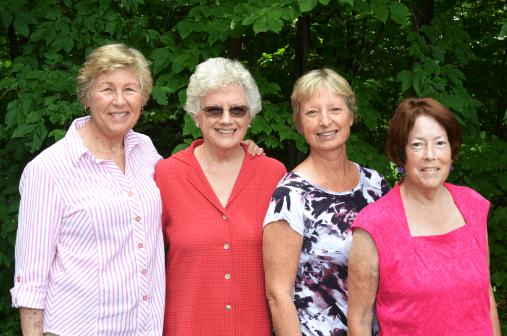 Four older women standing shoulder to shoulder outside by trees.
