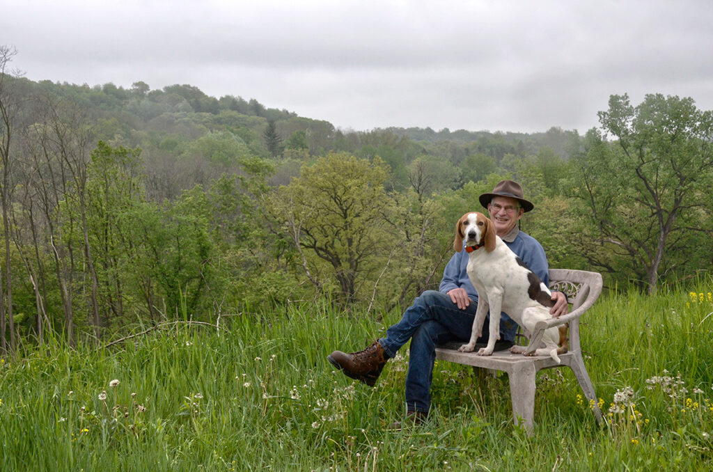 An older man sits on a bench with a dog in an open field with trees behind.