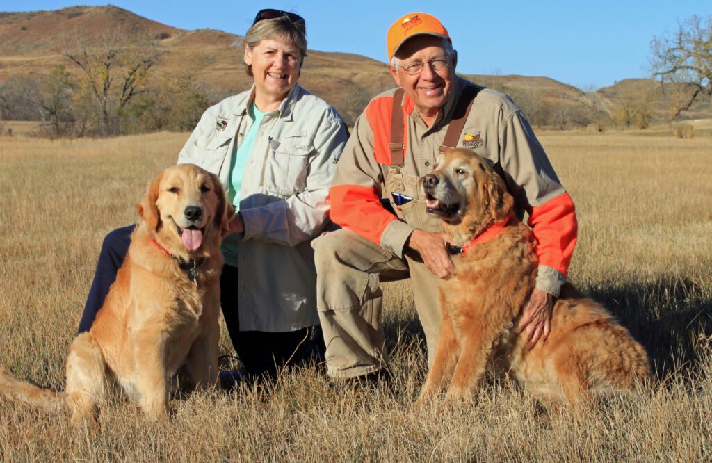An older woman and man kneeling with their two golden retrievers in a field in the fall.