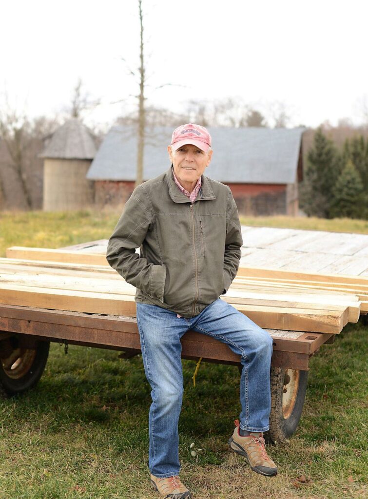 An older man in a beige jacket and baseball hat sits on a trailer in front of an old barn.