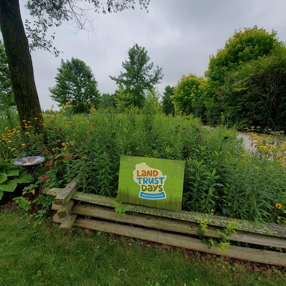 A green Land Trust Days yard sign propped up on a railing in front of lush green grass and trees.