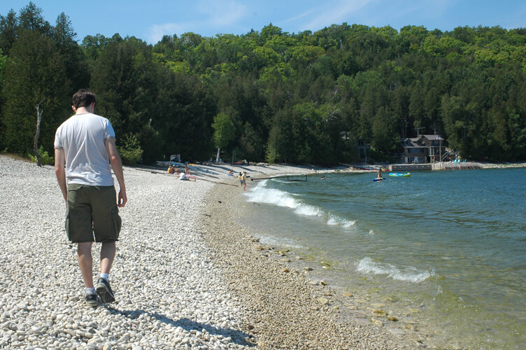 A man in a white t-shirt and shorts walking up a stony beach.