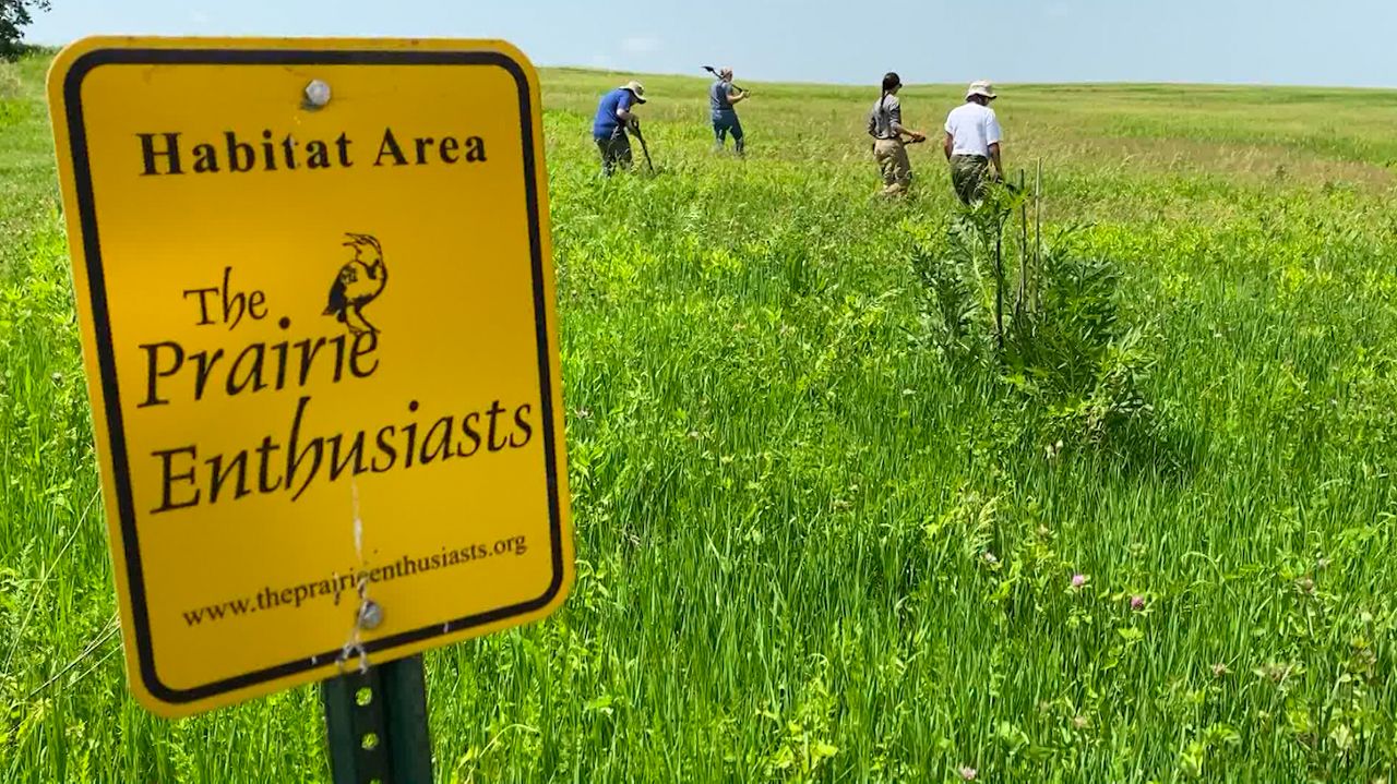 Yellow sign that says "Habitat Area: The Prairie Enthusiasts" with several people working in a field