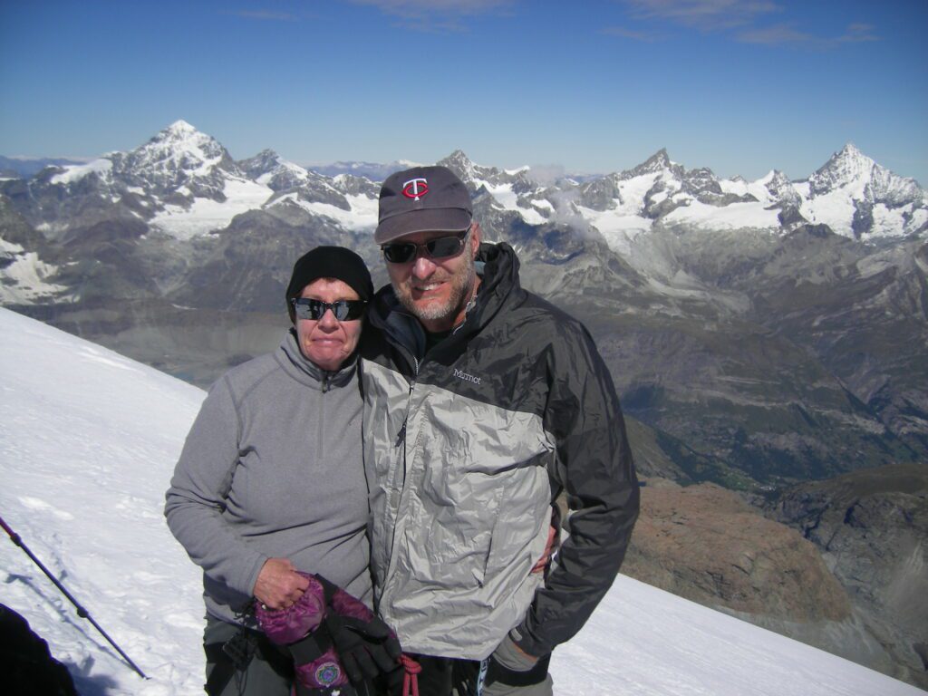 A man and woman standing side by side on top of a snowy mountain.