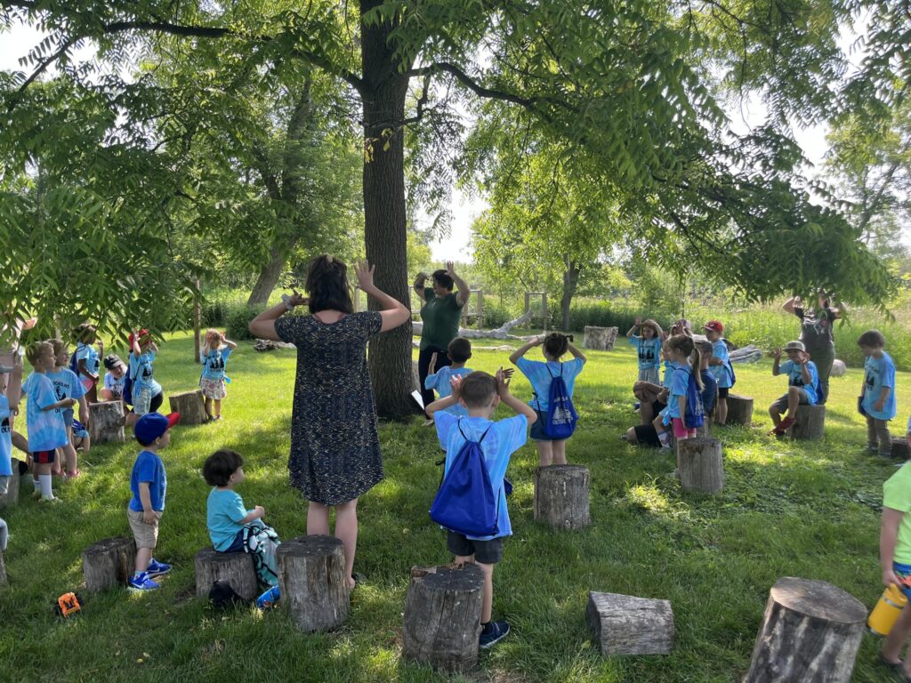 About a dozen kids playing a game with two adults around a tree in a grassy area with rock sitting stones