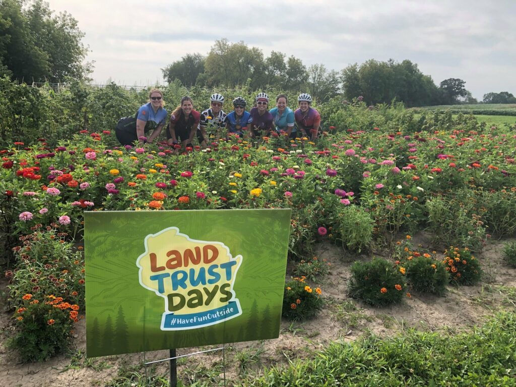 A green yard sign that says Land Trust Days in front of a group of a half dozen people kneeling in a flower bed