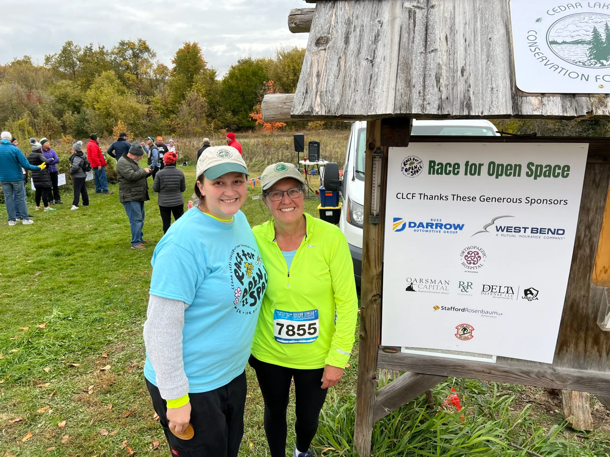 Two women in running gear standing in front of a sign that says "Race for Open Space" and a small crowd in the background.