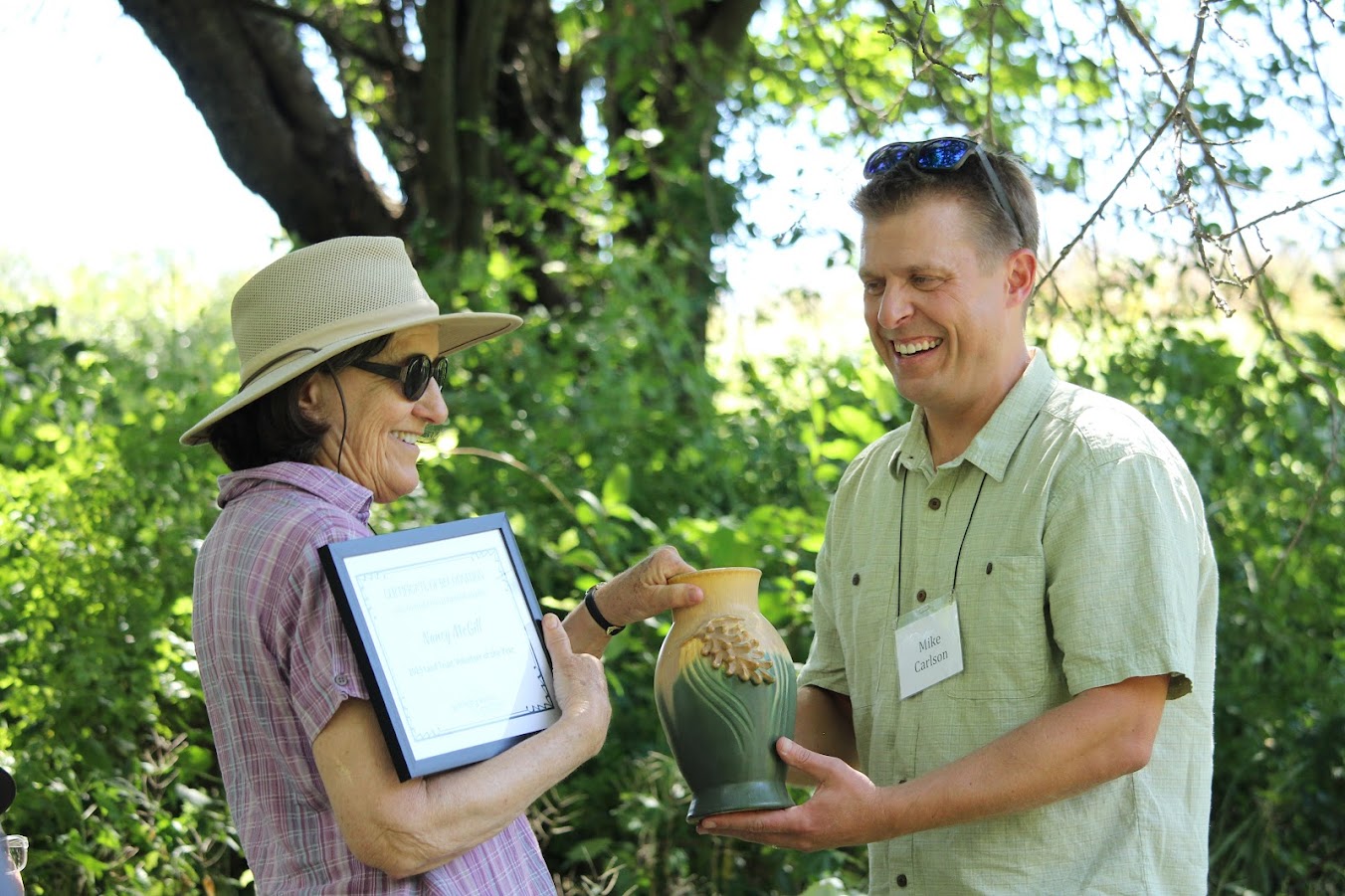 A woman receiving a framed certificate and ceramic vase form a man in an outdoor setting.