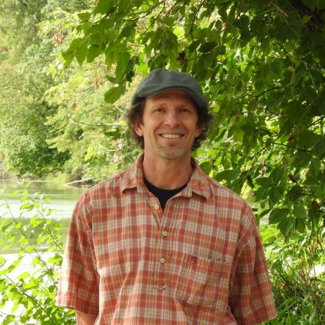 A middle-aged man in an orange shirt and brown hat standing outside by a tree.