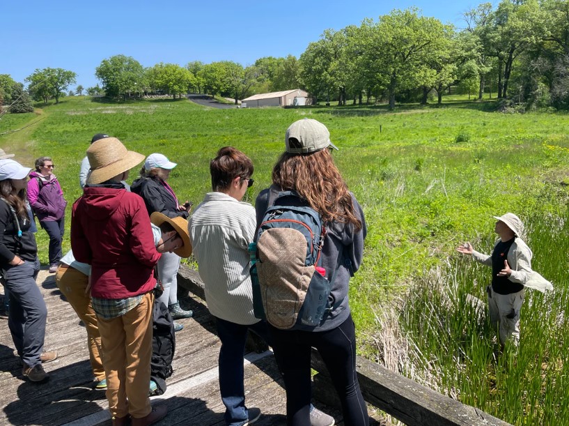 Several people standing on a wooden platform looking at a person talking below in a field.