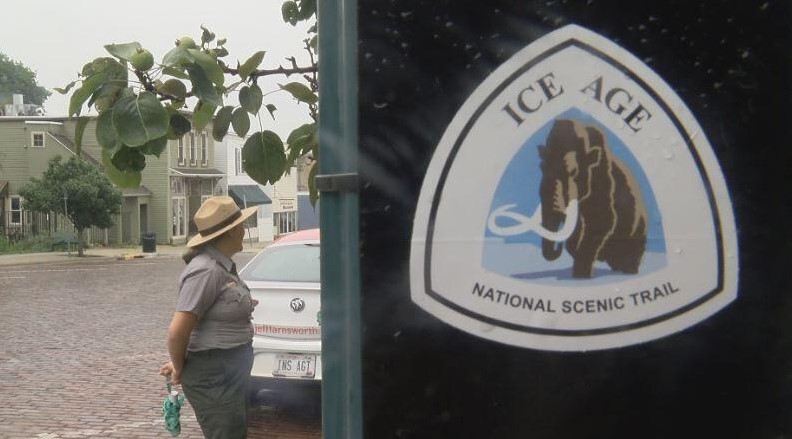 The Ice Age National Scenic Trail logo on a door with a female park ranger standing in the street nearby.
