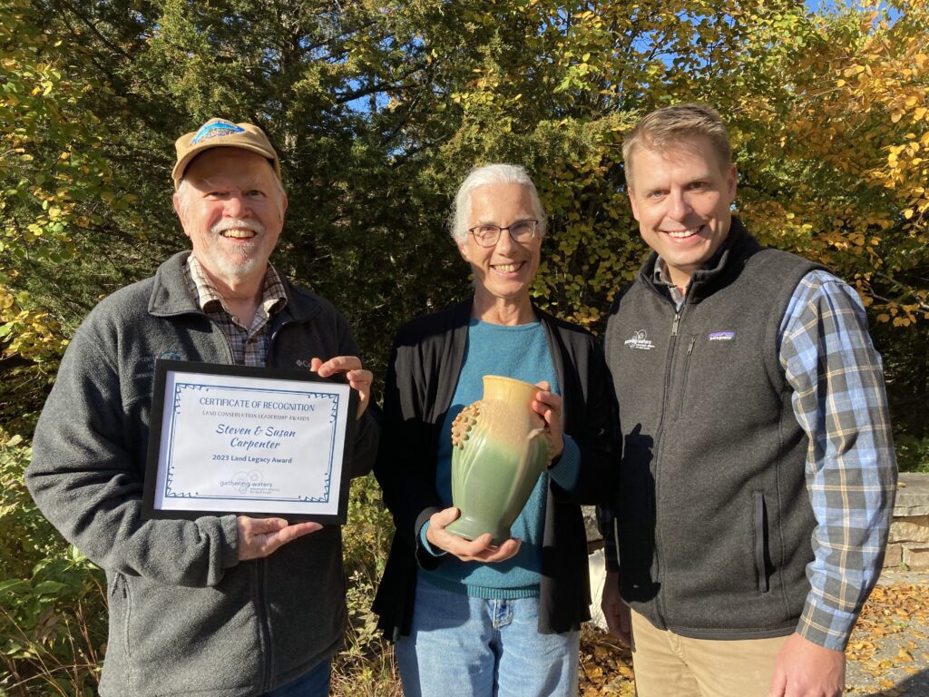Three people standing outside on a sunny day displaying an award certificate and ceramic vase.