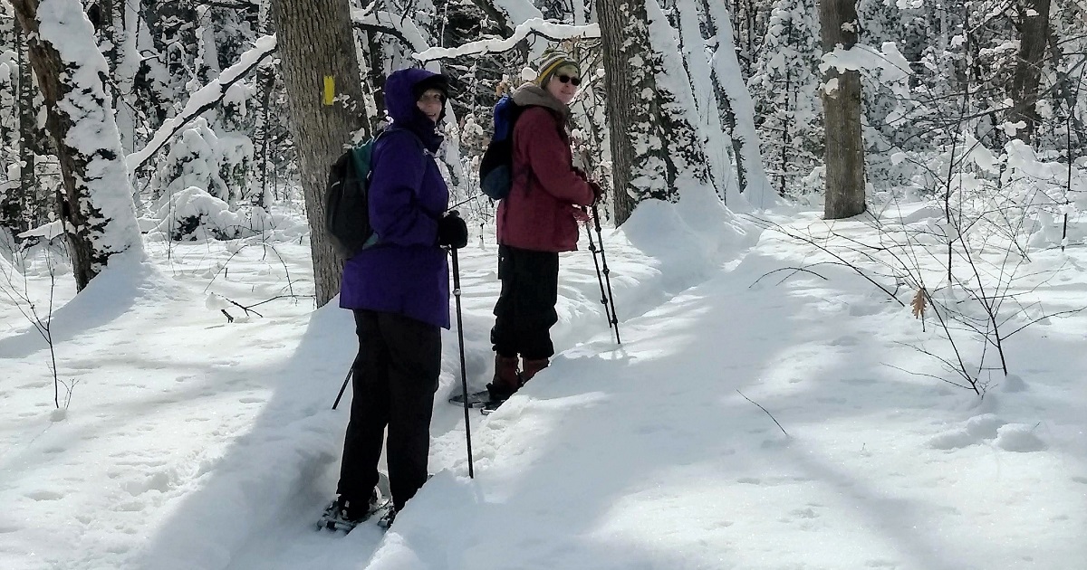 Two women snowshoeing on a trail with a yellow Ice Age Trail marker.