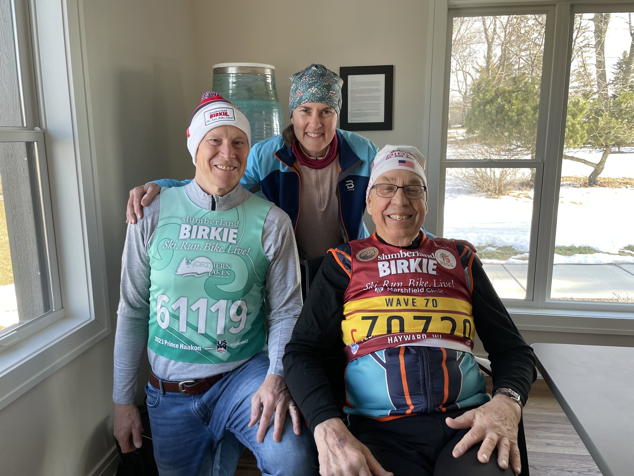 Three people posing in a living room wearing ski bibs and hats.