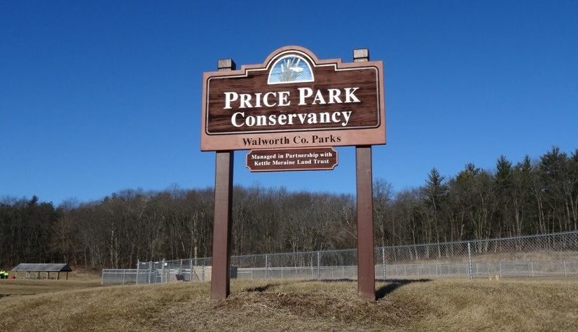 A wooden park sign stands against a clear blue sky above a fenced-in park area. A forest of tall pines span across the middle of the photo in the background. The sign states "Price Park Conservancy, Walworth County Parks, Managed in Partnership with Kettle Moraine Land Trust"