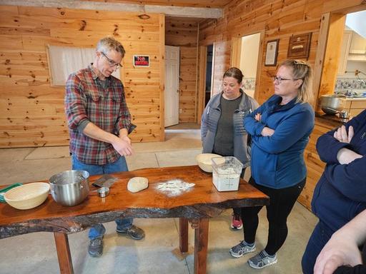 A photo of a group of individuals standing around a hand carved wooden table watching a baker through the process of making sourdough bread.