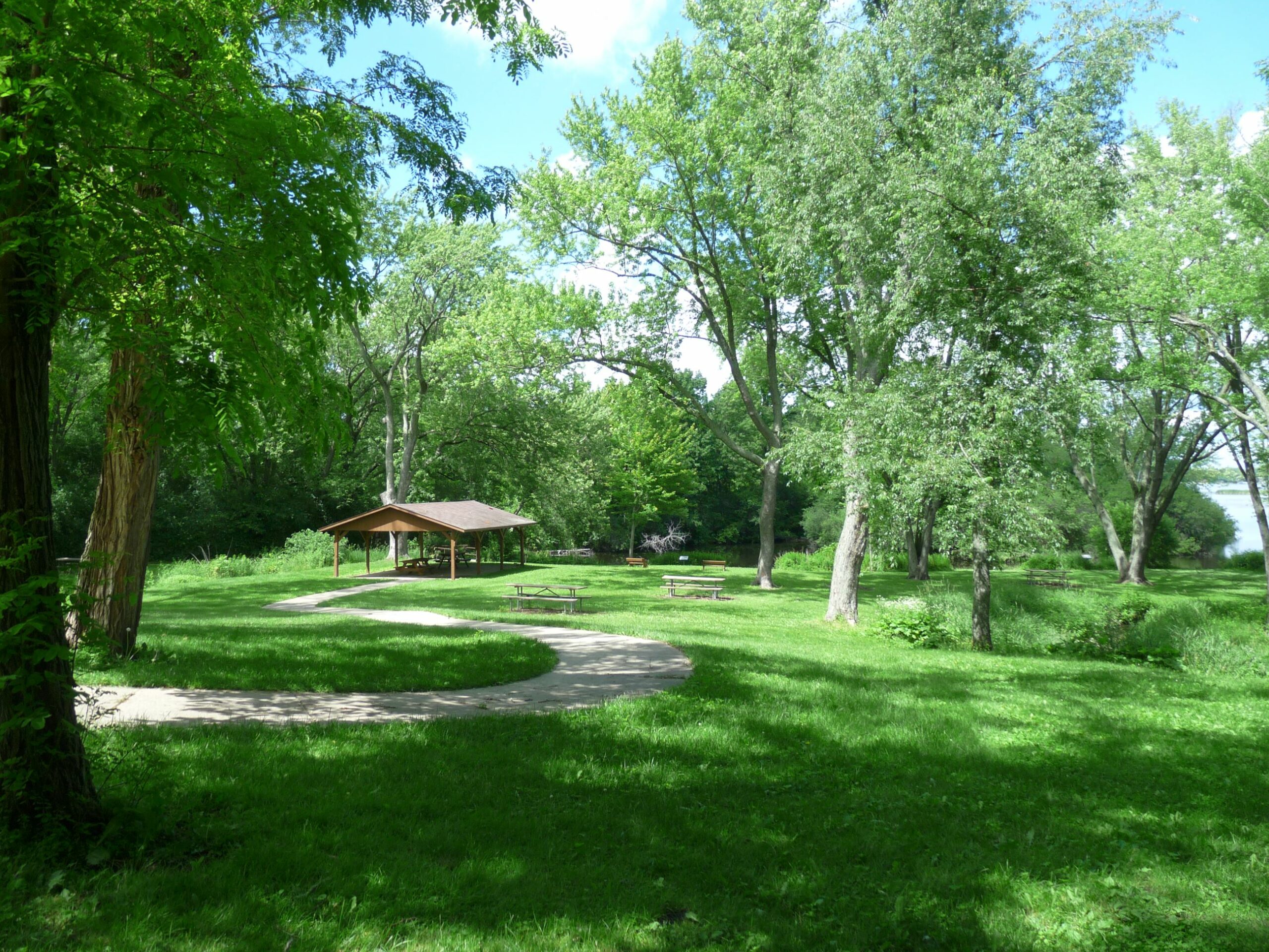 A landscape photo of a grassy park area. A winding sidewalk passes by picnic benches and ends at a park shelter in the middle of a grassy field.