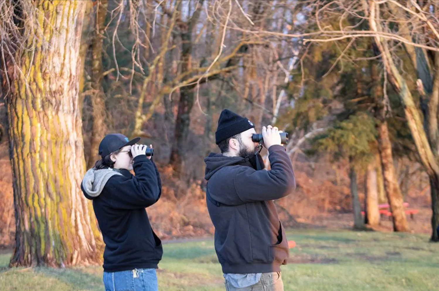 A side profile of two individuals, dressed for colder weather, looking out of binoculars in search of local birds.