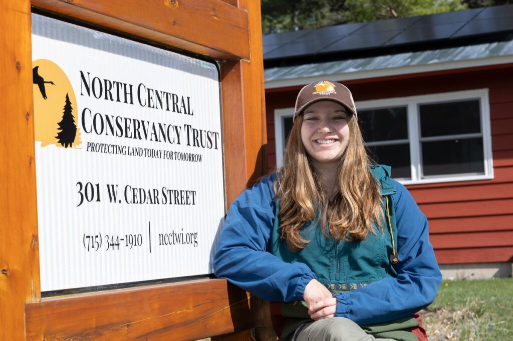 A young woman in blue jacket and brown baseball hat standing next to a sign for North Central Conservancy Trust.