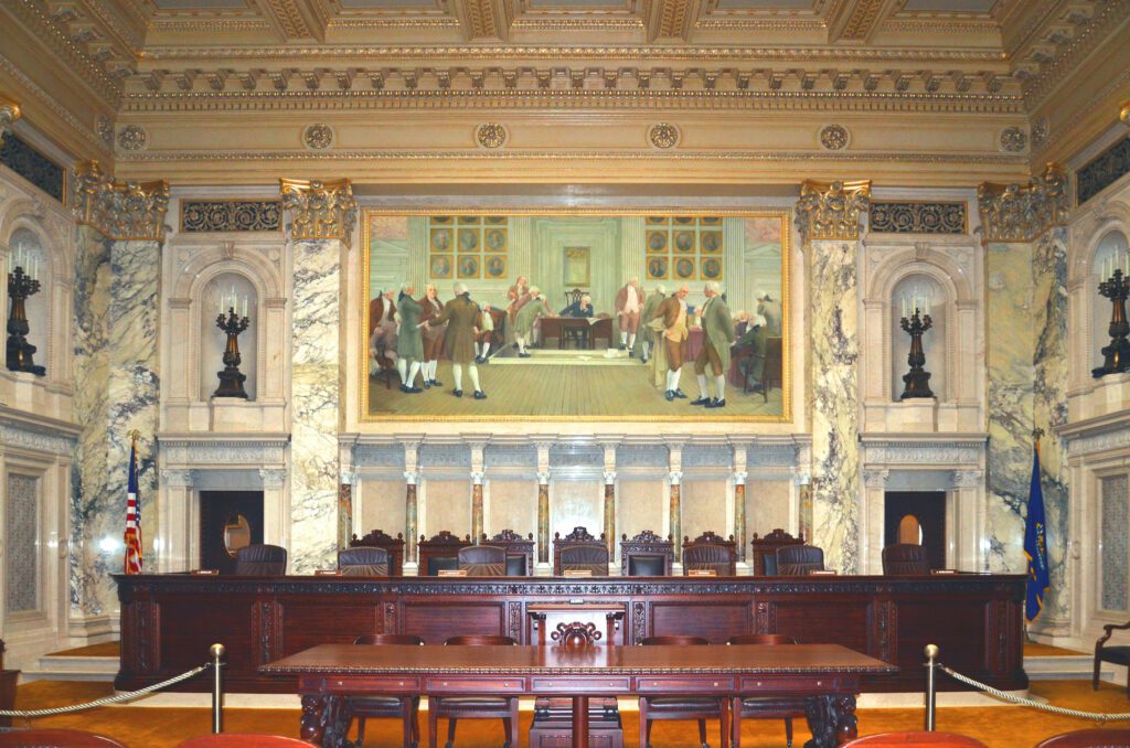 The Wisconsin Supreme Court Chamber with an elaborate wood seating area, large framed painting, and marble columns.