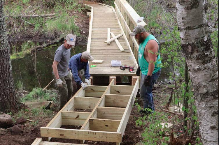 Three people constructing a wood bridge over a creek.