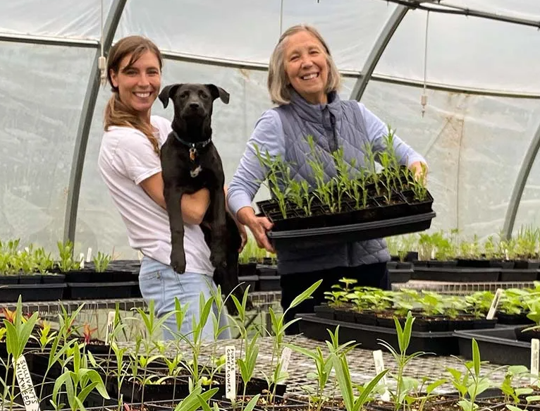 Two smiling women stand in a greenhouse. One woman is holding a black lab dog and the other is holding a tray of purple coneflower sprouts.