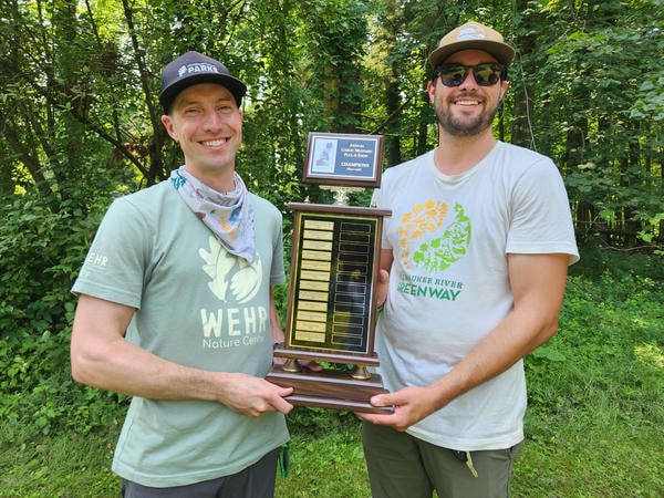 Two men in t-shirts standing outside holding a trophy.