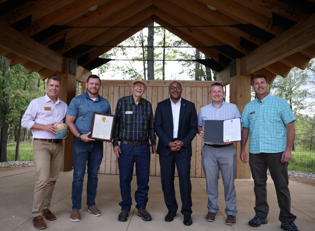 Six men standing in a row in an outdoor parks shelter posing for a photo after an award celebration.