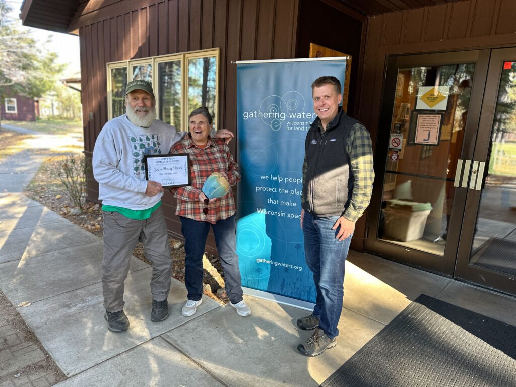 Three adults standing outside by a large blue banner, smiling and holding an award certificate and ceramic vase.