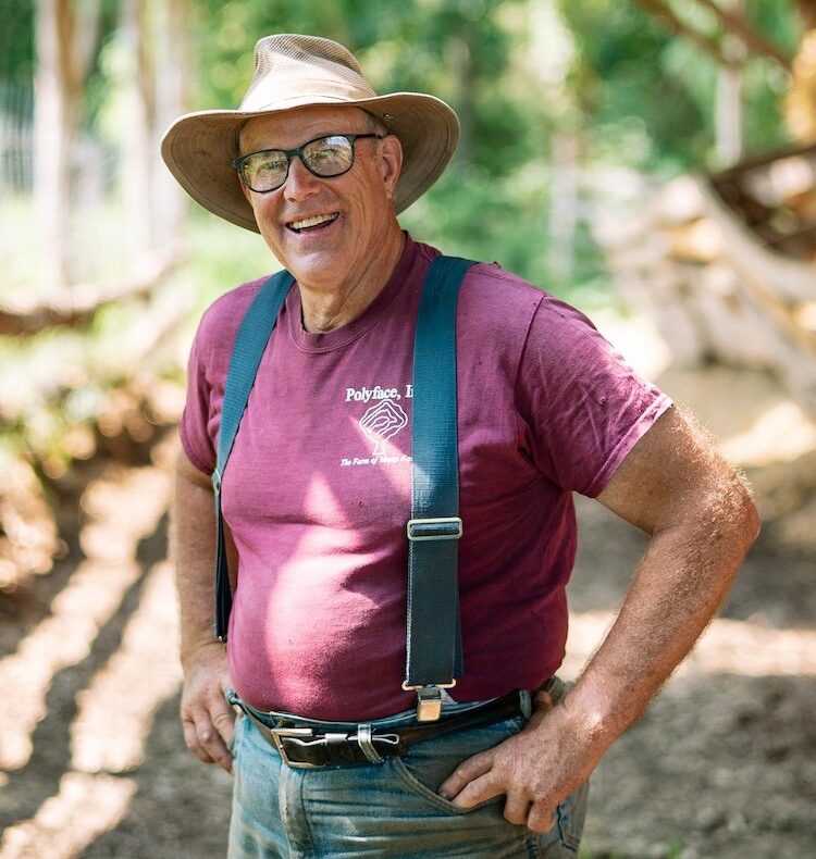 An older white man with maroon shirt and overalls standing outside.